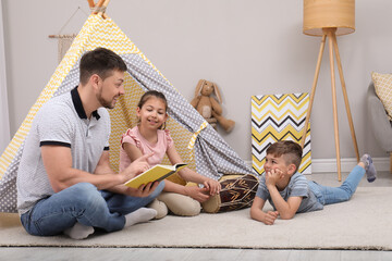 Father reading book to children near toy wigwam at home © New Africa