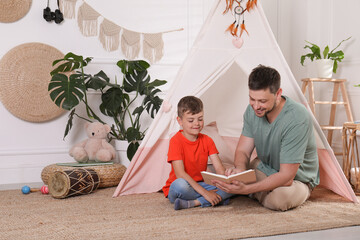 Father and son reading book near toy wigwam at home © New Africa