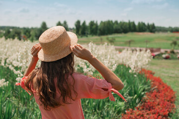 Woman in hat near flower bed. girl in pink dress, in straw hat stands with her back facing the sun in floral white and red field, holding the hat summer