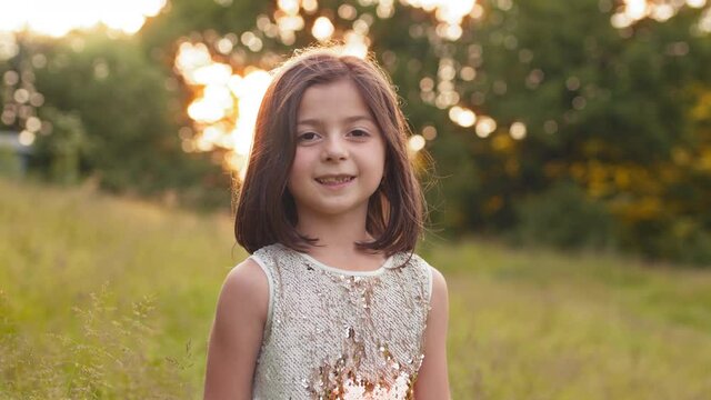 Portrait of pretty little girl with short brown hair posing at green garden with beautiful sunrise behind. Concept of happiness and childhood. Portrait of pretty girl with brown hair posing at garden
