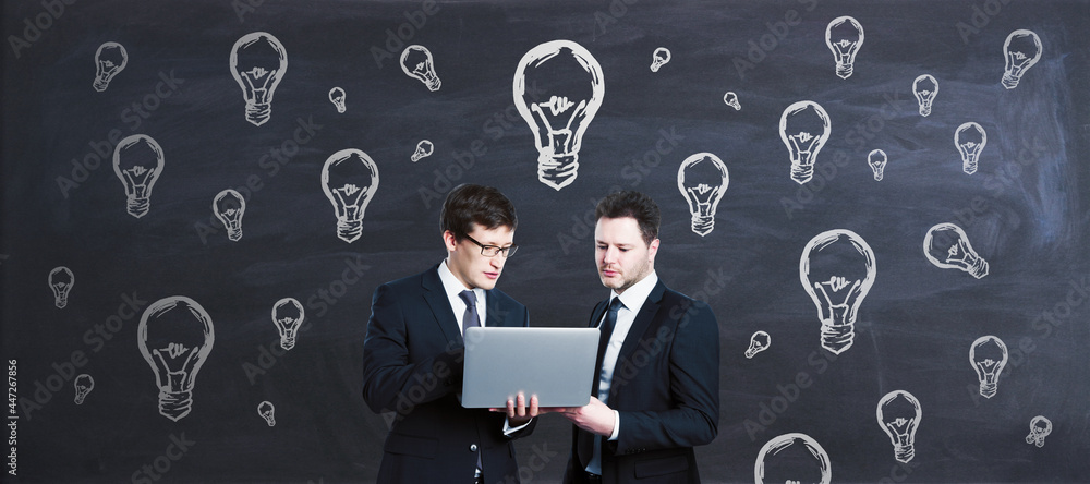 Poster two young european businessmen using laptop with abstract light bulb sketch on blackboard wall backg