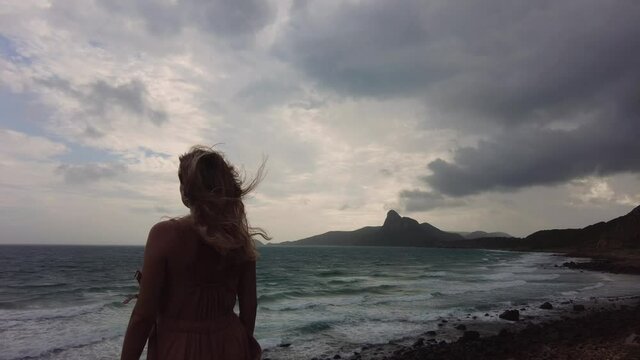 A girl is standing in front of the spectacular view of the ocean and some mountains on the horizon. Windy weather. Waves and white foam are scuttering along a rocky beach. Southeast Asia. Vietnam