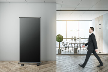 Businessman walking in modern coworking office interior with empty black banner, city view, equipment and furniture, law and legal concept. Mock up.