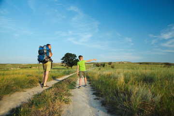 A young dad with a backpack and his little son travel among filds and hills, and boy is looking something by the hand and points to the sky into the distance, standing on the big road