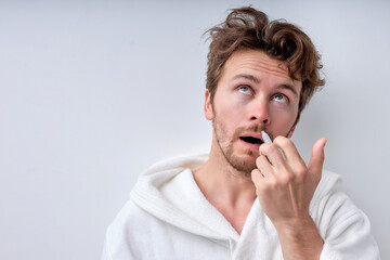 Portrait of attractive guy with nasal spray using nose drops over background, concept of treatment for allergies or common cold, winter style. bearded male in bathrobe suffering from disease