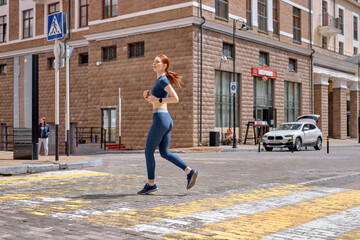 Fototapeta na wymiar Young woman running on road in city. Lifestyle morning run. Fitness and healthy lifestyle concept. Side view portrait on redhead lady in blue sportswear engaged in sport, jogging outdoors