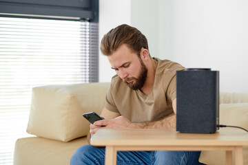 Young man turning on wireless smart column with the use of smartphone. Concept of a smart home wireless devices. Concept of smart home and voice command control