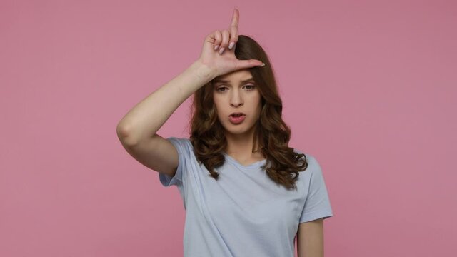 Adorable disappointed teenager girl in blue T-shirt holding fingers near forehead showing loser sign with sarcasm, unhappy because of crisis. Indoor studio shot isolated over pink background.