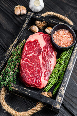Raw prime rib eye beef meat steak in a butcher wooden tray with herbs. Black wooden background. Top...