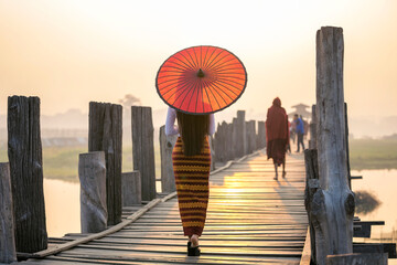 A young Burmese woman wearing traditional clothes with red umbrella at U Bein teak bridge in...