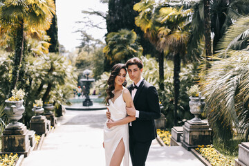 A beautiful married young couple in love in elegant wedding clothes walking posing and hugging on a romantic date in a green old park among plants and palm trees in summer in nature, selective focus