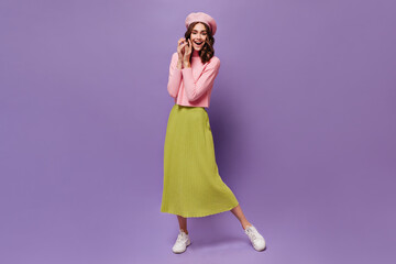 Cheerful curly woman in midi skirt and beret smiles on purple background. Attractive girl moves on isolated backdrop.