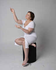 Full length portrait of young plus sized woman with short brunette hair,  wearing  tight white body con dress,   sitting pose on chair with gestural hands with light studio background.