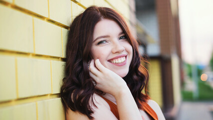 Beautiful young woman is having fun outdoors on a yellow brick wall background. Wonderful girl with brown hair smiling at camera. Smiles sincerely