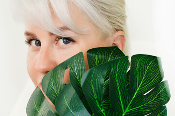 The face and eyes of a young blonde woman hid behind green monstera leaves. The concept of natural...