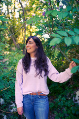 Happy and casual looking young woman portrait. She is smiling looking to her side standing in nature, surrounded of trees and purple flowers.