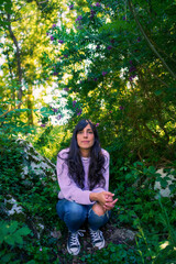 Hispanic latina woman looking at camera, sitting on some rocks surrounded by trees in nature. She wears casual clothes, jeans and purple sweater.