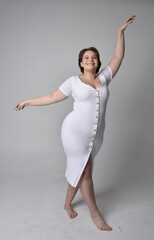 Full length portrait of young plus sized woman with short brunette hair,  wearing a tight white body con dress,   standing walking  posewith light studio background.