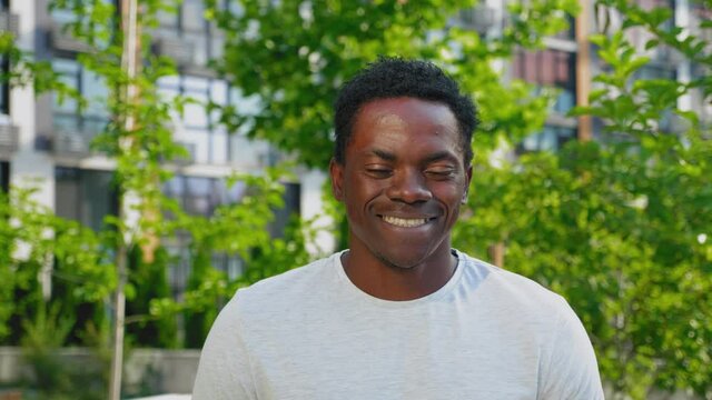 Close up smiling afro-american man looks camera on background modern building and trees in summer. Portrait positive African male wears gray t-shirt stands in park