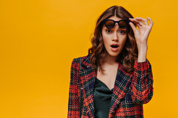 Emotional lady in striped outfit looking in surprise into camera on isolated backdrop. Curly girl with black sunglasses posing on yellow background..