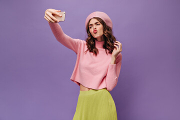 Obraz na płótnie Canvas Attractive curly woman in green skirt, pink beret and pink sweater blows kiss, takes selfie and holds phone on purple background.
