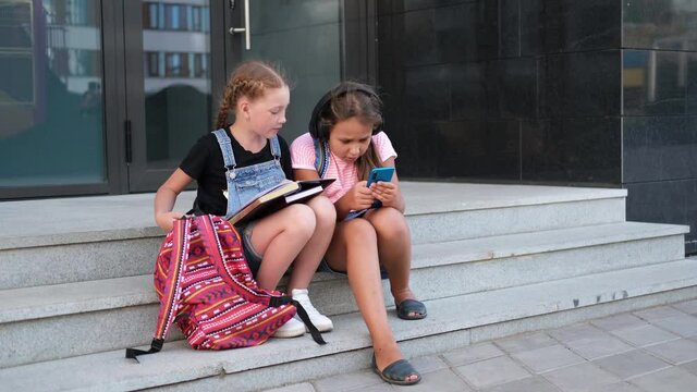  Two girls with backpack sit and read book with phone
