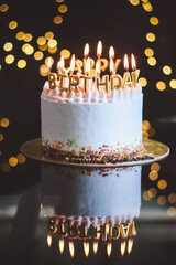 Birthday cake with candles, garland with bright bokeh lights on the background. The white cake is...