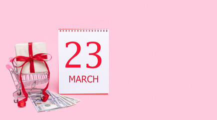 A gift box in a shopping trolley, dollars and a calendar with the date of 23 march on a pink background.