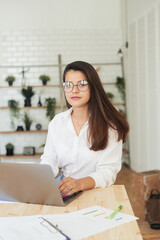 Thoughtful young woman in smart casual wear working while sitting in the office. Portrait of a young woman working at a laptop. Small business and business