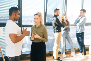 Obraz premium Side view of African American male posing with young woman colleague looking at camera, selective focus. Group of coworkers spending time together chatting and discussing new projects on background.