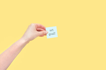 The hand holds a sticky note with inscription Set goal