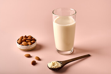 Almond milk in glass, flour in wooden brown spoon and nut kernels on pink powder background. Selective focus. Copy space