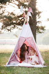 Beautiful mother with her little daughter sitting near wigwam in the park. Spending time together, outside, on vacation, outdoors. © Andriy Medvediuk