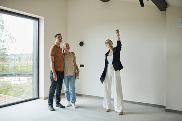 Full length portrait of female real estate agent giving apartment tour to young couple buying new...