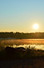 Late summer sunrise on a small pond. A mist floats above the water. The rising sun gilds the landscape with its light. There is an old boat on the beach.