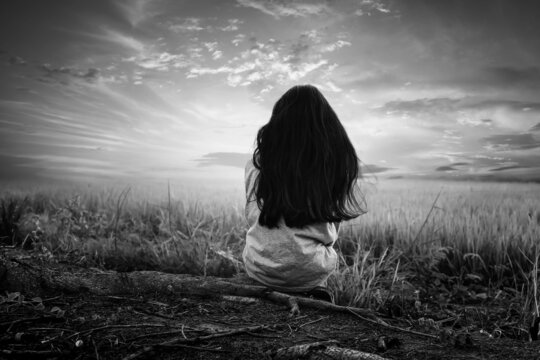A black and white picture of a young girl sitting in the middle of a rice field watching the sun go down alone.