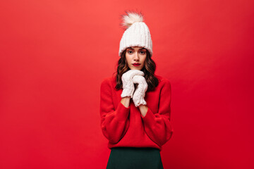 Curly woman in red sweater, knitted hat and mittens looks into camera. Lady in warm winter outfit poses on isolated bright background.