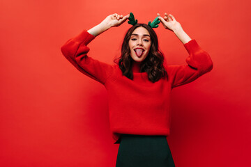 Happy girl in red sweater showing tongues on isolated backdrop. Joyful lady with curly hair posing on bright background..