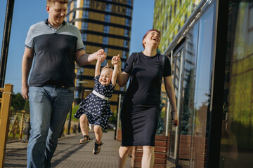 caucasian family couple walking with baby girl lifting baby by hands. Woman is happily laughing