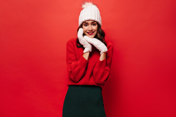 Cool cute woman in warm knitted hat, mittens and sweater smiles. Charming girl poses on red isolated background.