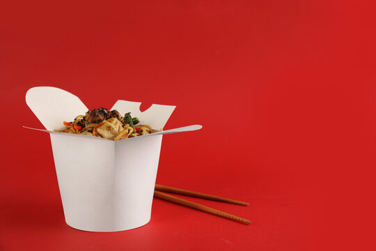 Box of wok noodles with vegetables, meat and chopsticks on red background. Space for text