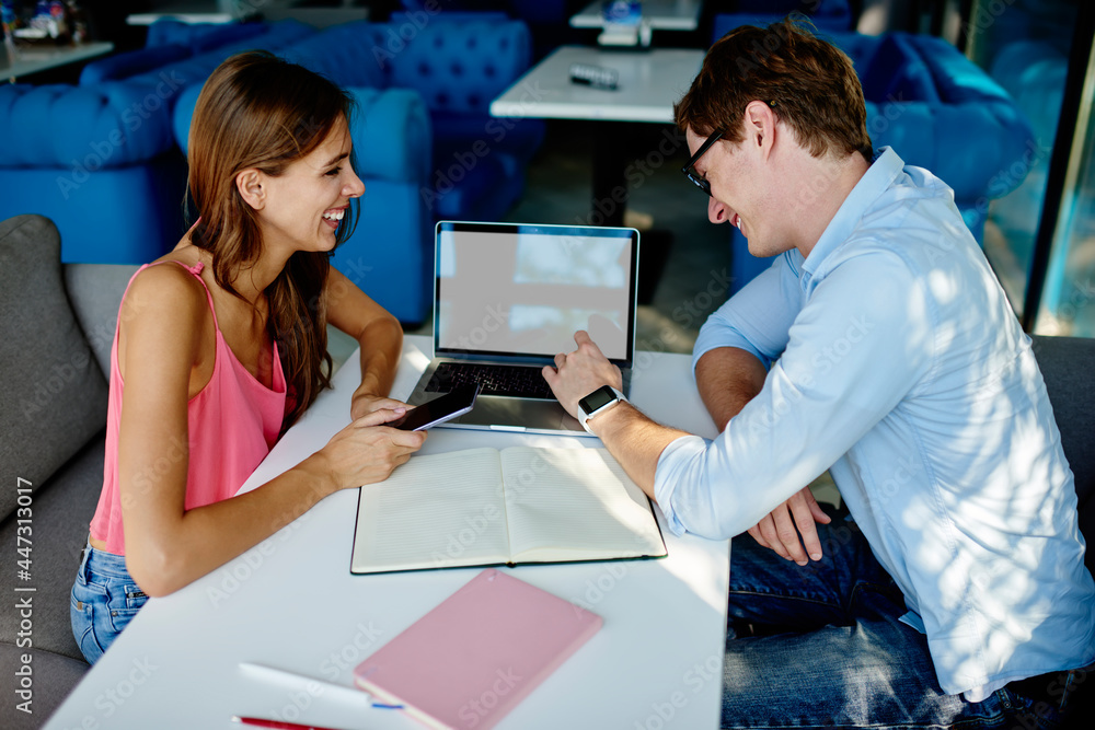 Poster cheerful male and female students laughing at table desktop with blank laptop computer enjoying coll