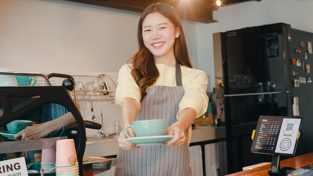 Portrait young Asian lady barista waitress holding coffee cup feeling happy at urban cafe. Asia small business owner girl in apron relax toothy smile looking to camera stand at counter in coffee shop.