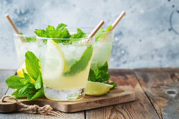 Two glass with lemonade or mojito cocktail with lemon and mint, cold refreshing drink or beverage with ice on rustic blue background. Copy space