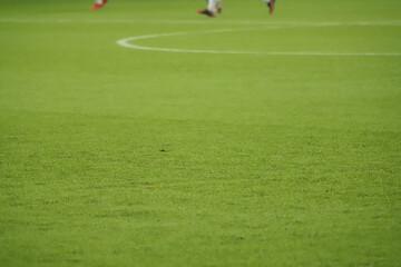 Synthetic green grass of a football field, highlighted by its texture, in the upper third, the players' feet
