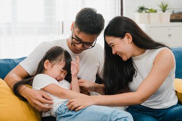 Happy cheerful Asian family dad, mom and daughter having fun cuddling playing on sofa while...