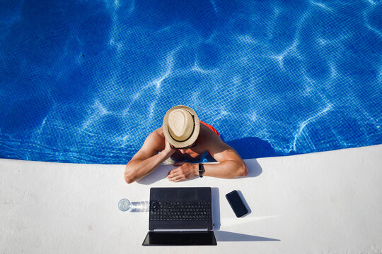 bird view of remote online working digital nomad man on workation with hat &  laptop at a white table standing in a sunny turquoise water pool