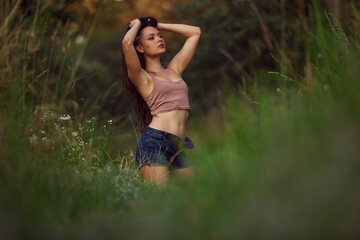 Naklejka na ściany i meble Sexy woman posing in a field in the grass rays of the sunset. Lifestyle on nature dressed in jeans shorts and beige top in golden hour. Romantic autumn mood, long tall grass in the meadow