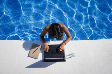 bird view of remote online working digital nomad woman in bikini with long black hair and laptop on a white table standing in a sunny blue water pool on Workation
