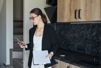 Graphic portrait of female real estate agent using smartphone while standing by kitchen in house...